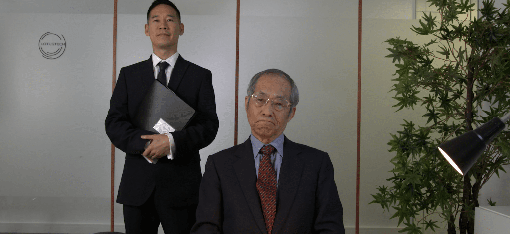 Man sitting at desk with plant to the right of him and a man standing holding some folders on his left.