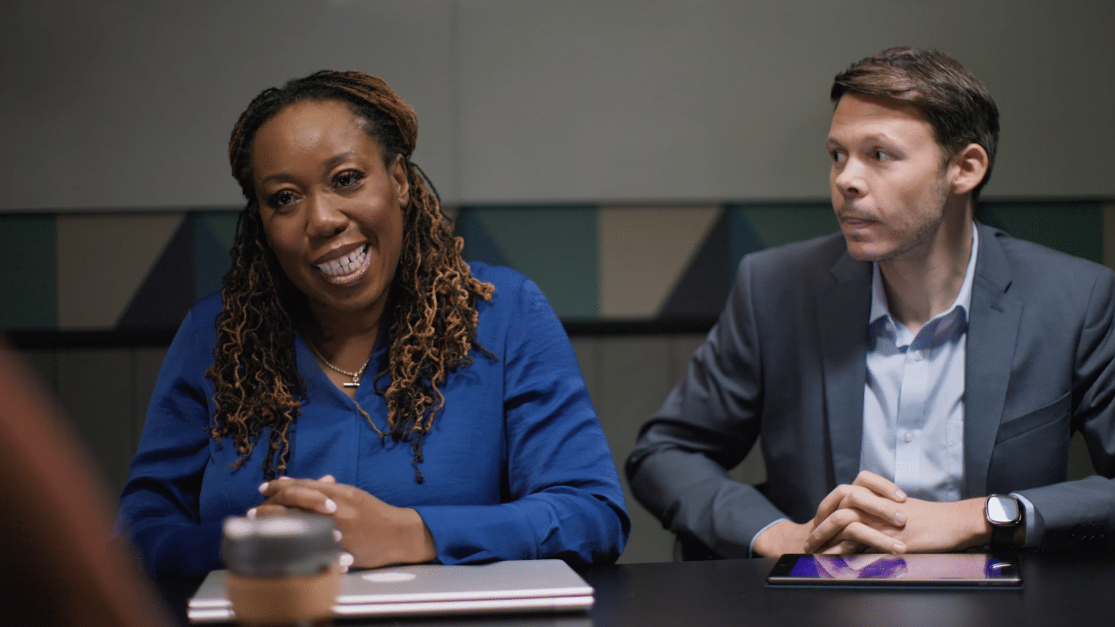 Man in suit and woman in blue top sitting at desk. Woman is smiling as man looks over at her