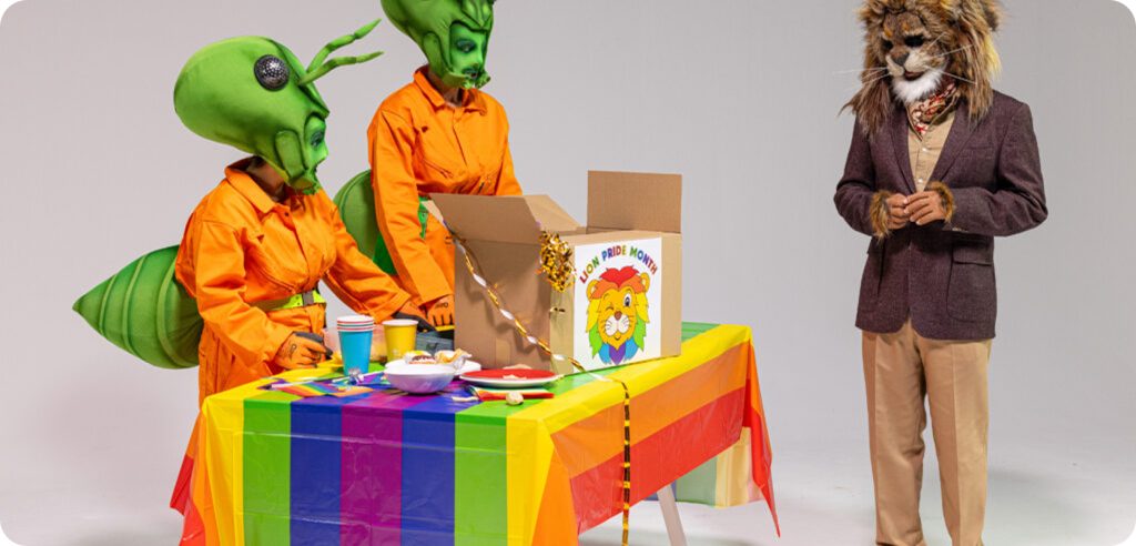 A man dressed as a lion talking to two women dressed as ants standing at a table with a Pride flag on it.